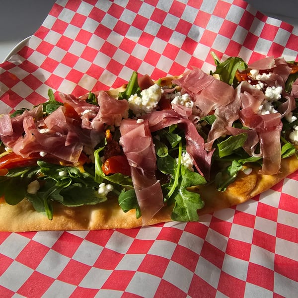 Flatbread with prosciutto, arugula, goat cheese, and sun-dried tomatoes on red and white checkered paper.