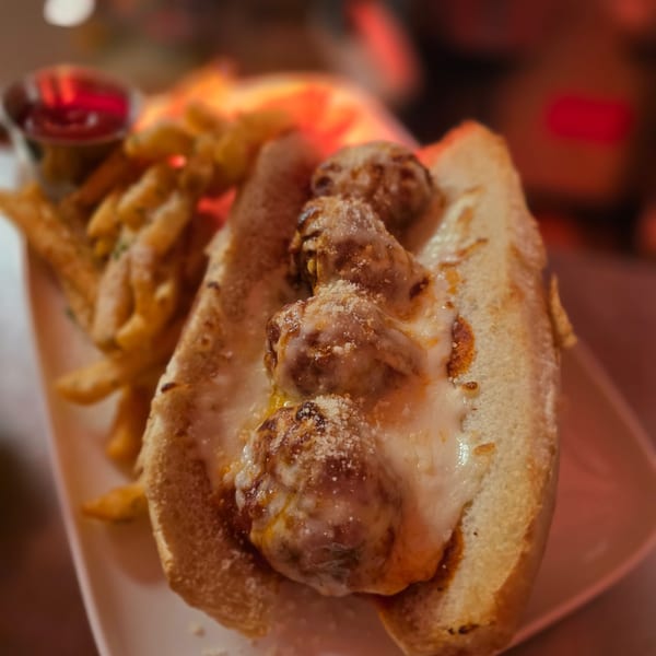 Meatball sub sandwich with melted cheese and fries on a white plate, with a red dipping sauce.
