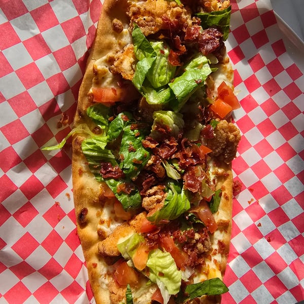 A crispy flatbread pizza topped with fried chicken, bacon, lettuce, and tomatoes on checkered paper.